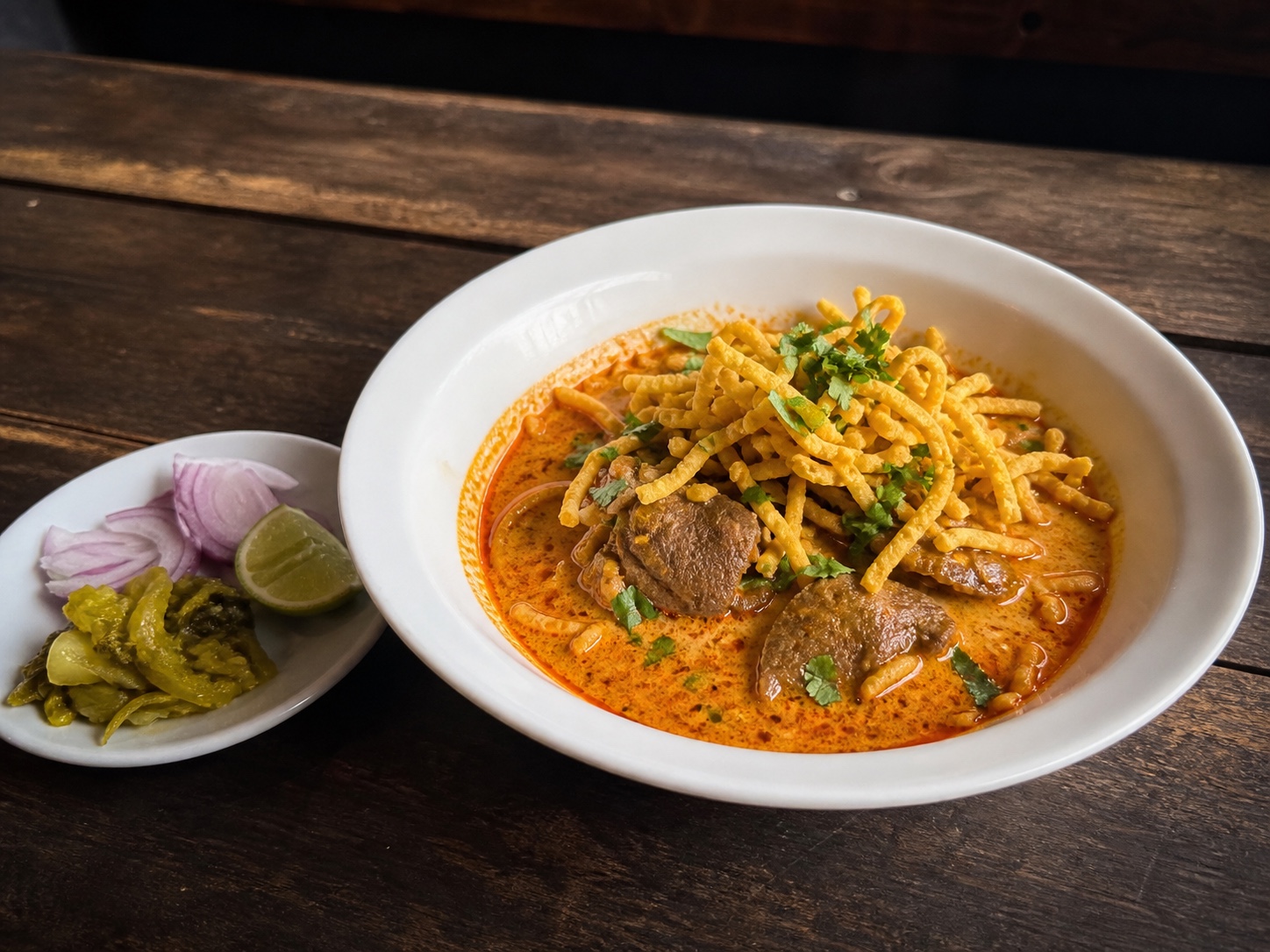 ชามข้าวซอยที่ร้านข้าวซอยกลอยใจ หมู่บ้านแม่กำปอง, เนื้อตุ๋นในน้ำซุปกะทิแกงแดง เส้นหมี่กรอบโรยหน้า ผักชีสด เครื่องเคียงหอมแดงซอย มะนาว และผักกาดดอง บนโต๊ะไม้